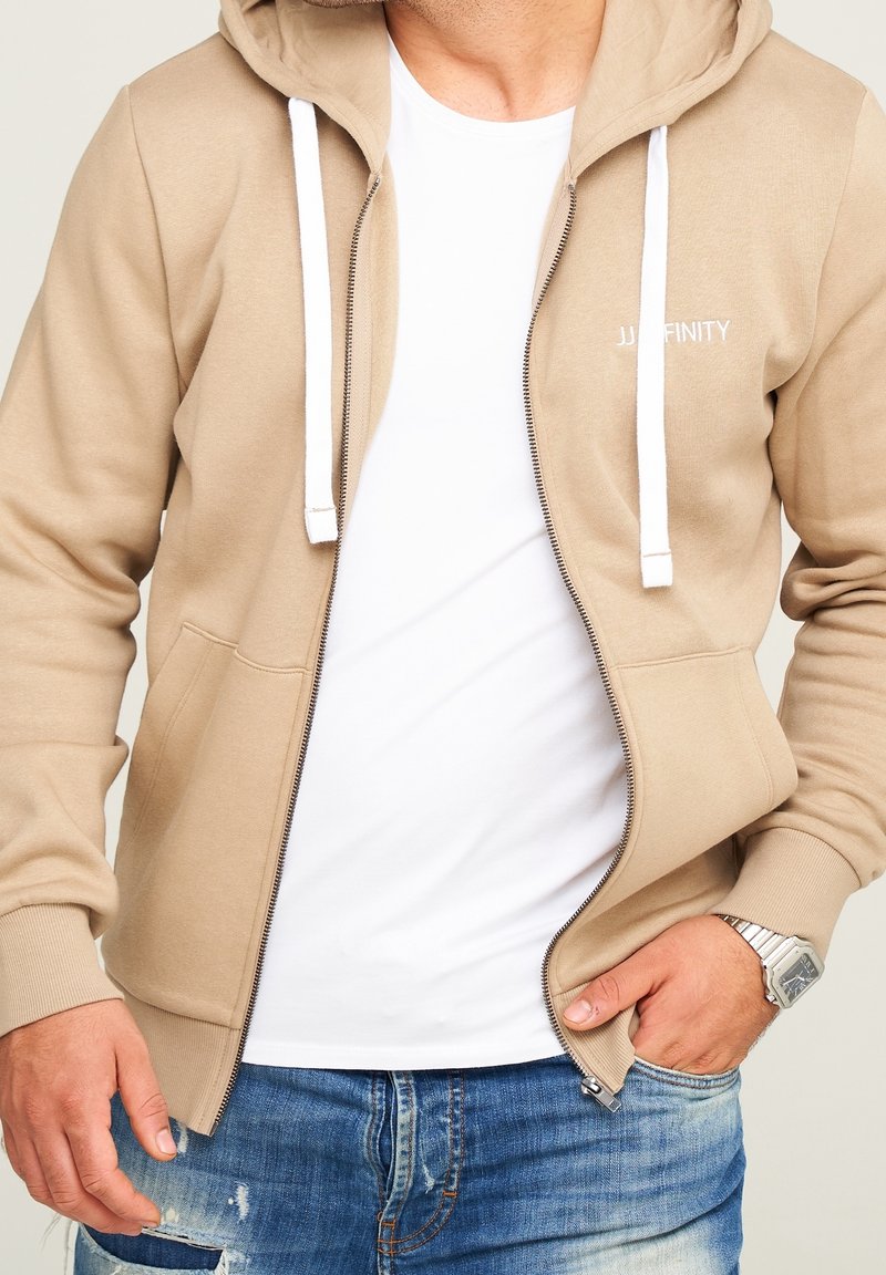 Homme portant un sweat à capuche beige zippé par-dessus un t-shirt blanc et un jean bleu, avec une main dans la poche et une montre-bracelet argentée.