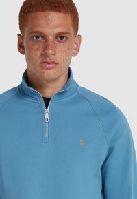 Felpa zip-up azzurro chiaro realizzata in tessuto morbido, con colletto alto, zip argentata e un piccolo logo arancione sul petto.