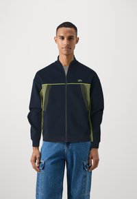 Casaco de fecho éclair azul-marinho com detalhes verdes e painéis laterais transparentes. Apresenta uma gola alta e punhos canelados. O modelo usa calças cargo azuis.