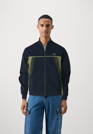 Veste zippée marine avec des accents verts et des panneaux latéraux transparents. Dotée d'un col montant et de poignets côtelés. Le modèle porte un jean cargo bleu.