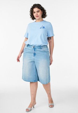 Kvinde med krøllet hår iført en lyseblå T-shirt med et lille fugledesign, lange denimshorts og lyseblå sandaler med hæl.