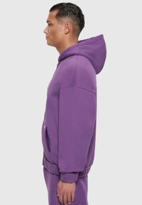 Lila Kapuzenpullover mit einer Fronttasche, gerippten Bündchen und einer Kordelzugkapuze. Aus weichem Material mit lockerer Passform, seitlich dargestellt.