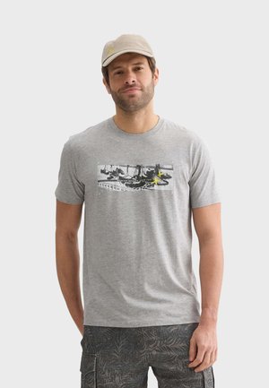 Camiseta de algodón gris con un estampado gráfico de zapatillas, mangas cortas y cuello redondo. El hombre también lleva una gorra beige.