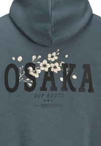 Kapuzenpulli in gedämpftem Teal, mit fettem schwarzen Text "OSAKA" und floralen Grafiken in Weiß. Weiches Material mit lässiger Passform.