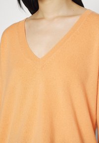 V-kaulusneule pehmeässä oranssissa neulosmateriaalissa, jossa on rento malli. Kankaan pinta on sileä ja hihat ovat hieman laskeutuvat.