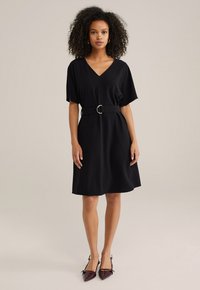 Robe noire à col en V avec des manches courtes et une ceinture à la taille dotée d'une boucle ronde. Tissu lisse avec une coupe décontractée et une longueur au genou.