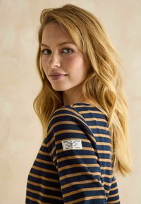 Jeune femme aux longs cheveux blonds portant une chemise rayée bleu marine et beige avec un petit écusson sur la manche supérieure, regardant par-dessus son épaule.