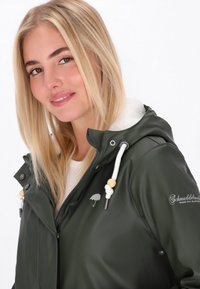 Tumši zaļš ūdensizturīgs apmetnis ar flīsa odere kapuci. Iezīmes ietver auklu detaļas, sudraba lietussarga logotipu un pogas.