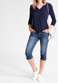 Top de manga larga azul marino con escote en V y bolsillos delanteros, combinado con pantalones capri de mezclilla. La modelo lleva zapatillas blancas y lleva un bolso cruzado con estampado.
