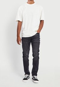 Uomo che indossa una maglietta bianca oversize a maniche corte, jeans neri slim fit scoloriti e sneaker bianco e nero, in piedi con una mano in tasca.