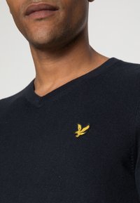 Maglione blu navy a scollo a V con maglia testurizzata; presenta un piccolo logo di uccello ricamato giallo sul petto a sinistra.