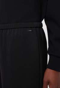 Gros plan d'un pantalon noir à taille élastique avec une petite étiquette métallique du logo Armani Exchange sur la taille, à côté de la main d'une personne dans une manche noire.