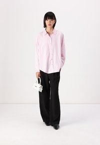 Camicia a righe rosa e bianche con maniche lunghe, abbinata a pantaloni larghi neri. Il modello tiene una piccola borsa bianca.