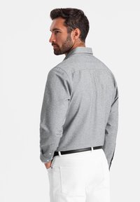 Chemise grise à manches longues avec une finition texturée, col boutonné et poignets simples. Portée avec un pantalon blanc, elle présente un détail de pli dans le dos.