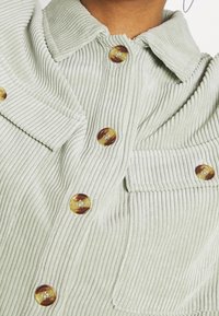 Ljusgrön kordflortsskjorta med framträdande vertikala ribbor, stora bruna knappar och krage, samt en fickdetalj på bröstet.