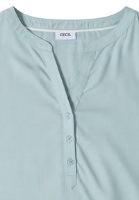 Blouse bleu clair avec col rond et patte de boutonnage à trois boutons, avec étiquette de marque "CECIL" à l'intérieur du col.