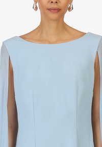 Robe bleu clair avec un décolleté carrée et des manches en cape fluides. Tissu lisse avec une texture subtile, complété par des boucles d'oreilles en argent.