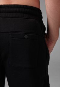 Black sweatpants featuring a soft texture, elastic waistband, and a rear pocket with a black snap closure.