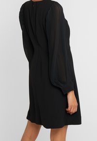 Schwarzes Kleid mit plissierten, halbtransparenten Ärmeln. Hat ein tailliertes Oberteil, eine schmale Taille und geknöpfte Manschetten, die über dem Knie enden.