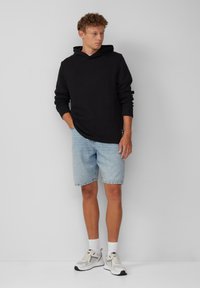 Schwarzer Hoodie mit Kängurutasche, kombiniert mit hellblauen Denim-Shorts. Das Model trägt graue und weiße Sneaker mit weißen Socken.