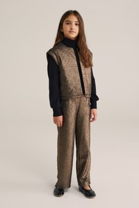 Texturerad bronsväst och matchande byxor i glansigt tyg, bärs över en svart polo. Svarta skor fulländar outfiten.