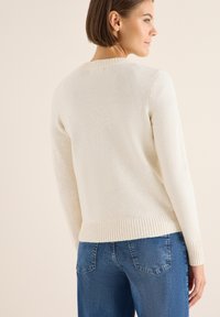 Krämfärgad tröja med ribbat avslut och ärmslut, med en slät yta och en klassisk rund halsringning. Bärs med blå jeans.