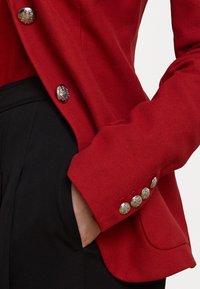 Červený texturovaný blazer s dekorativními stříbrnými knoflíky na manžetách, v kombinaci s černými kalhotami. Tkanina má strukturovaný, tkaný vzhled.