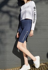 Bílý dlouhý rukáv, cropped top a tmavě modré cyklistické šortky, obé s značkou "Columbia". Modelka má na sobě bílé platformové tenisky.