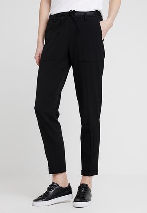 Persona que lleva pantalones negros con cordón y bolsillos delanteros, y zapatillas negras con suelas blancas, de pie frente a un fondo liso.