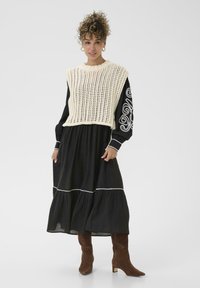 Chaleco de punto crema sobre un vestido negro con ribete blanco, que presenta mangas largas y un diseño en capas. Botas de tacón de ante marrón completan el look.