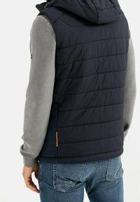 Námořnická bunda bez rukávů s kapucí, která má prošívanou texturu a šedé dlouhé rukávy. Obsahuje malou oranžovou etiketu blízko lemu.