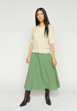 Kobieta o długich czarnych włosach ubrana w kremowy krótki sweter na guziki, zieloną spódnicę midi w kształcie litery A oraz brązowe, spiczaste botki, stojąca na jasnym tle.