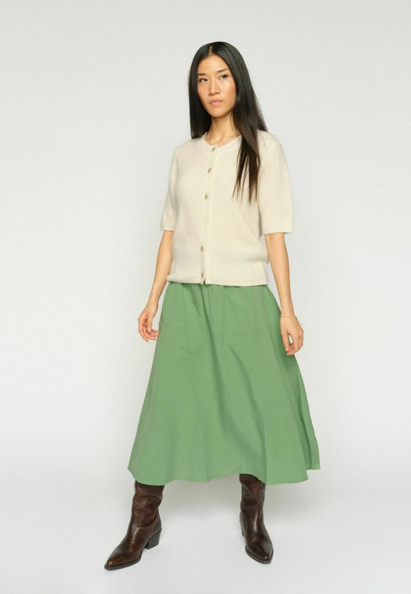 Hosszú fekete hajú nő krémszínű, gombos, rövid ujjú kardigánt, zöld A-vonalú midi szoknyát és barna hegyes orrú csizmát visel, világos háttér előtt áll.