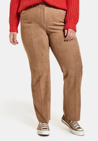 Kobieta w beżowych zamszowych spodniach w prostym kroju, czerwonym swetrze dzianym i beżowych trampkach z białymi sznurówkami.