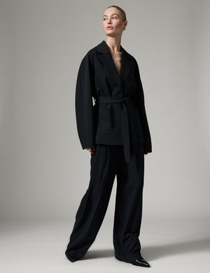 Kvinde står iført en oversized sort bælteblazer og vidde bukser med spidse sorte sko mod en ensfarvet grå baggrund.