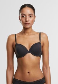 Femme avec une expression neutre portant un soutien-gorge noir à dentelle et un sous-vêtement assorti sur un fond gris uni.