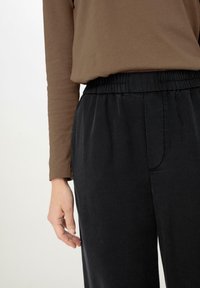 Bruine lange mouwen top gecombineerd met zwarte elastische broek met tailleband. De top is zacht, terwijl de broek een gladde textuur en subtiele zakdetails heeft.