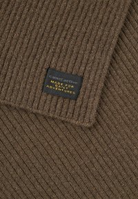 Bruine geribde gebreide sjaal met een label waarop "cameactive" staat en de tekst "MADE FOR DAILY ADVENTURES" in het geel.