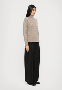 Frau, die einen taubfarbenen, kabelgestrickten Rollkragenpullover, weite schwarze Hosen und schwarze Schuhe trägt, vor einem schlichten weißen Hintergrund steht.