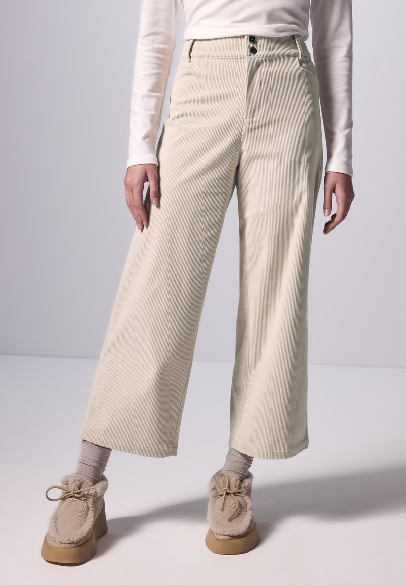 Ljust beige kordflorbyxor med vida ben, två framknappar och texturerade vertikala linjer. Bärs med grå strumpor och beige fluffiga skor.