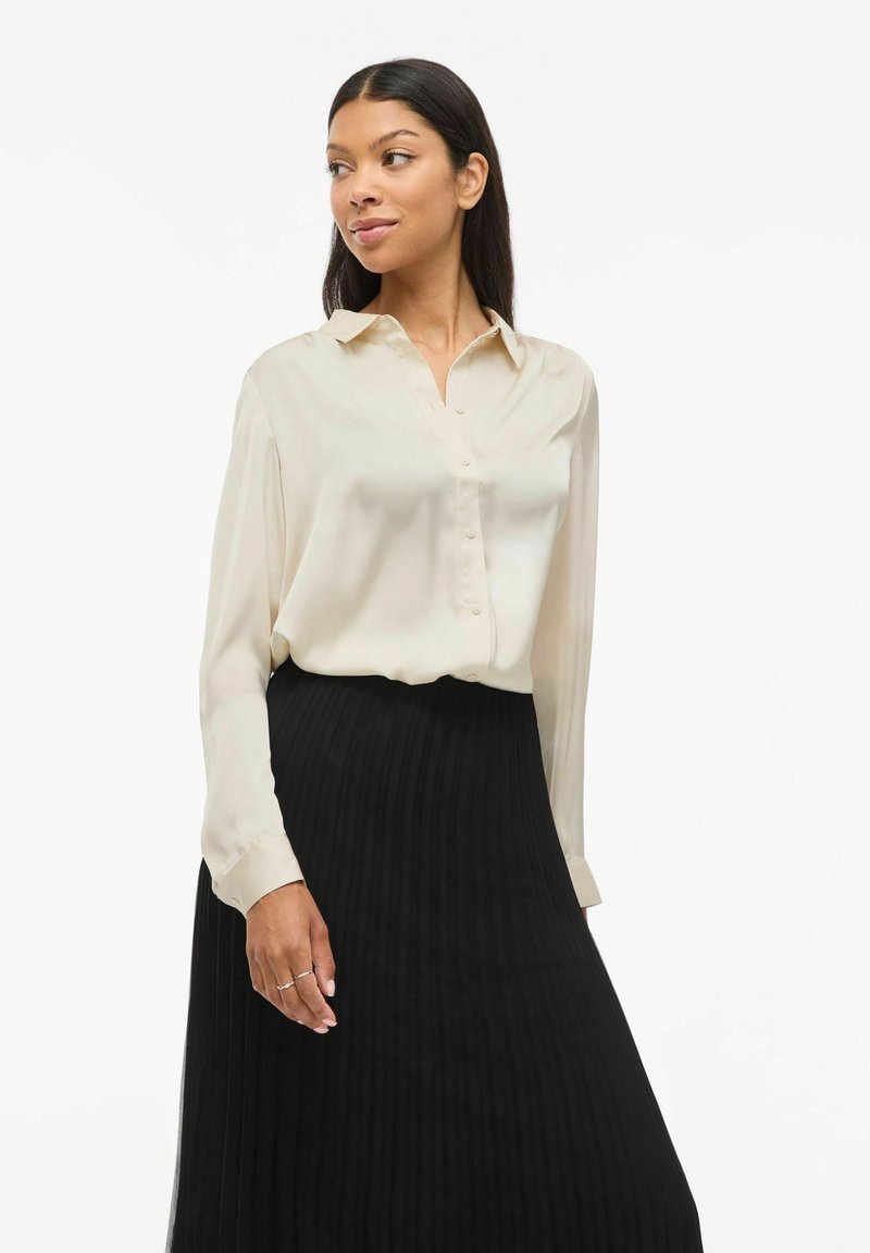 Crème blouse met een knoopsluiting aan de voorkant en lange mouwen, gecombineerd met een zwarte plissérok. De materialen lijken soepel en licht van gewicht.