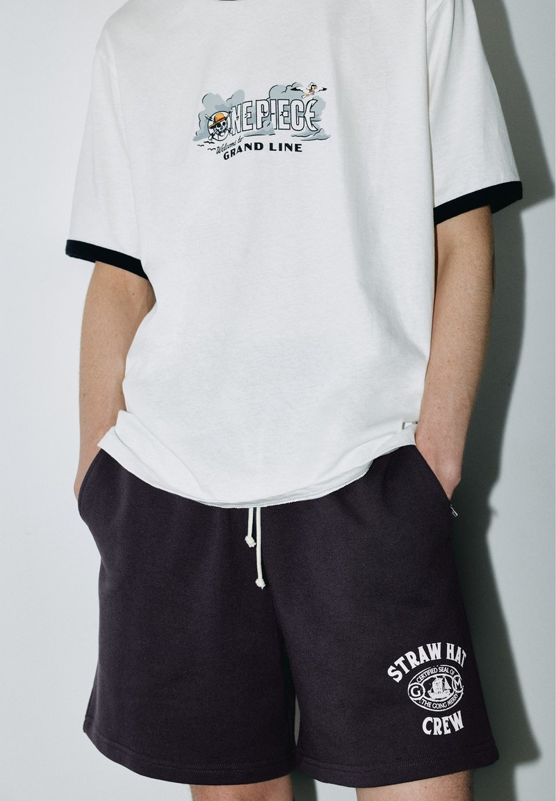 T-shirt blanc avec bordure noire présentant le logo One Piece Grand Line et short noir avec cordon de serrage et texte Straw Hat Crew sur la jambe gauche.