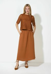 Braune Spitzenbluse mit kurzen Ärmeln, kombiniert mit einem fließenden braunen Rock. Der Rock verfügt über Seitentaschen und Schnürdetails am Saum. Braune Absätze.