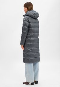 Long manteau matelassé gris avec capuche, présentant des sections matelassées horizontales. Le manteau est brillant et léger, associé à un jean bleu clair à jambes larges.