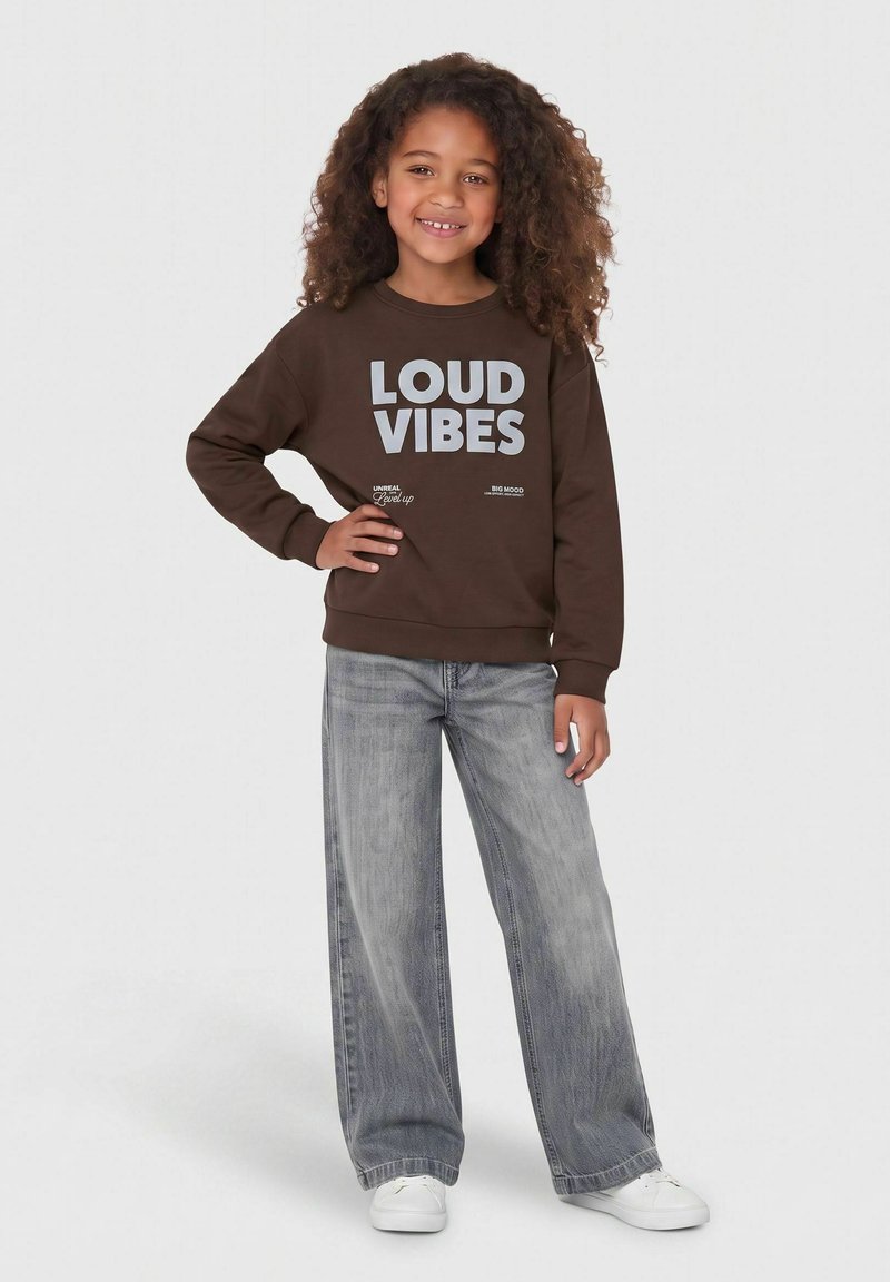 Fille souriante aux cheveux bouclés, debout, main sur la hanche, portant un sweat-shirt marron avec l'inscription "LOUD VIBES,", jean gris et chaussures blanches.
