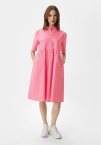 Abito di cotone rosa con maniche corte, colletto, apertura con bottoni, vita elasticizzata e tasche laterali; abbinato a sneakers bianche.