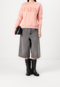 Sudadera rosa con "PARIS" en texto naranja bordado, combinada con culottes grises y botas negras hasta la rodilla, complementada con un bolso negro pequeño.