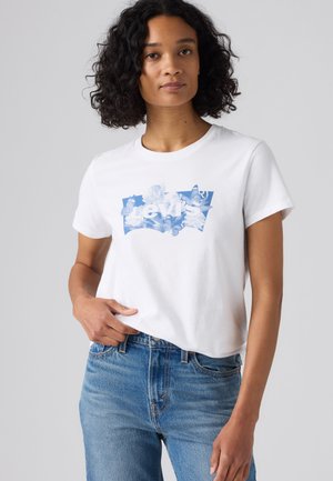 Femme aux cheveux bouclés portant un t-shirt blanc Levi's avec un logo floral bleu et un jean bleu, debout devant un fond gris clair uni.