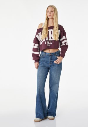 Kvinde med langt blond hår iført cropped vinrød sweatshirt med bare skuldre og teksten "Paris", højtaljede udvidede blå jeans og hvide sneakers.