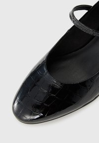 Chaussure Mary Jane en cuir verni noir avec une texture crocodile et une fine bride sur le dessus, vue en gros plan sous un angle.